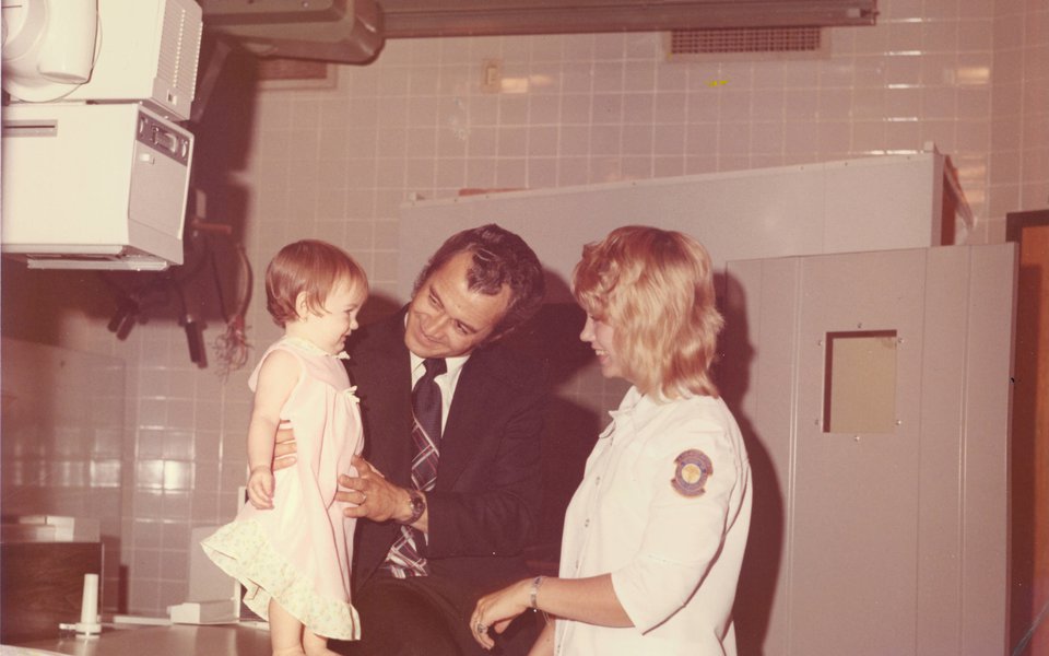 A historic photo of a small child smiling at a CoxHealth nurse while standing on a radiology table, circa 1975
