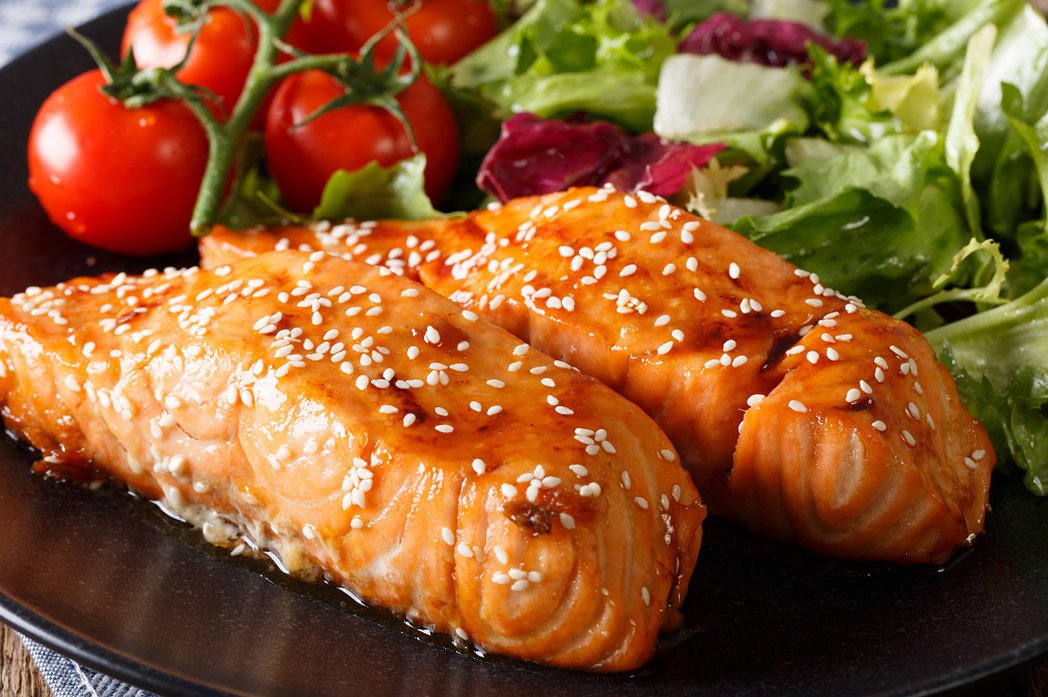 A close-up of glazed salmon on a black plate with fresh cherry tomatoes and salad.